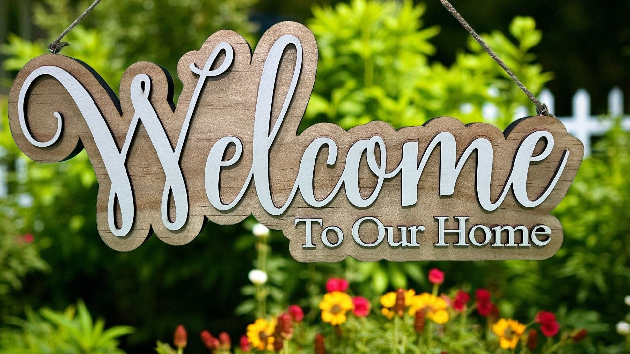 Wooden 'Welcome To Our Home' sign hanging in a garden with flowers and greenery.